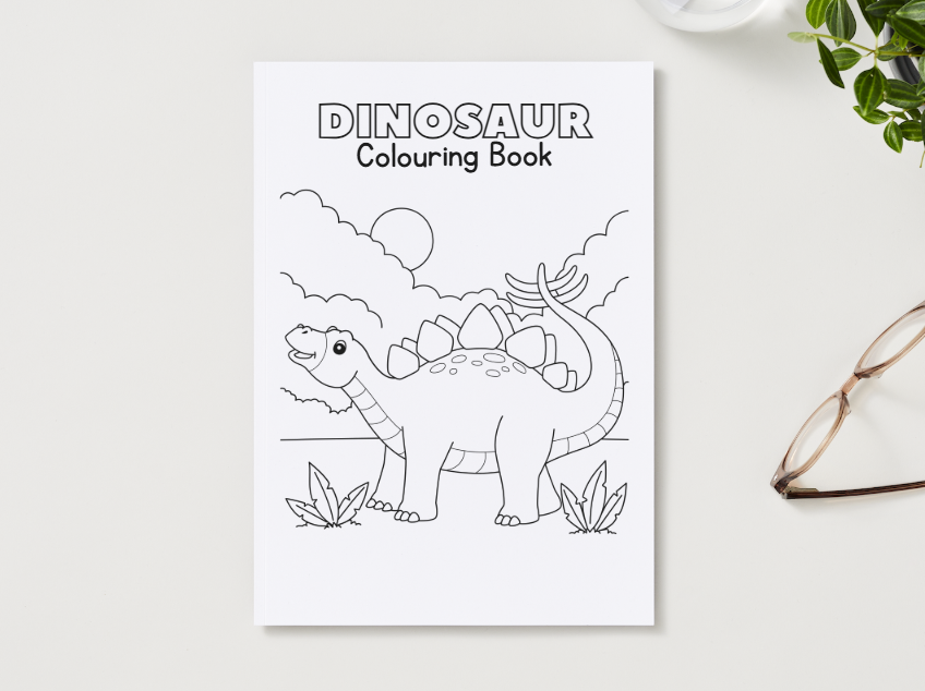 Dinosaur coloring book on a white surface with a plant and glasses in the background