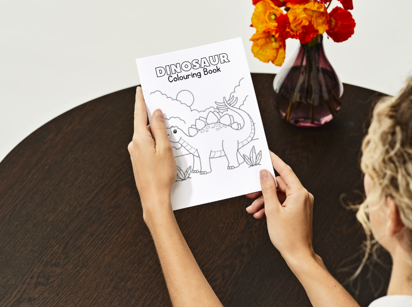 Person holding a dinosaur coloring book on a wooden table with flowers in the background