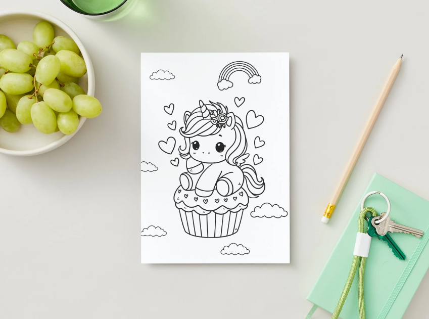 Coloring book page with a unicorn in a cupcake design on a table with grapes and stationery items.